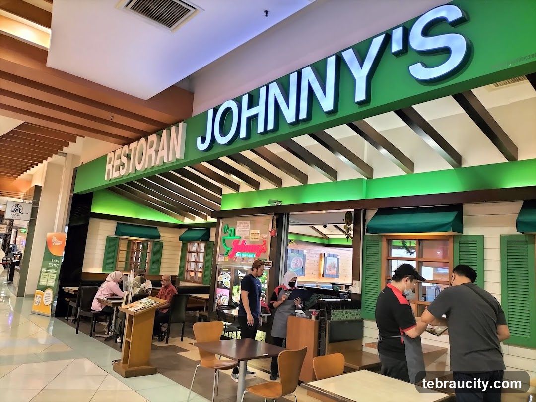 The exterior of a restaurant called Restoran Johnny's. The restaurant has a green facade with white siding and green shutters on the windows. A large green sign with white lettering that says "RESTORAN JOHNNY'S" is visible above the entrance. Several people are seated at tables inside and outside the restaurant.