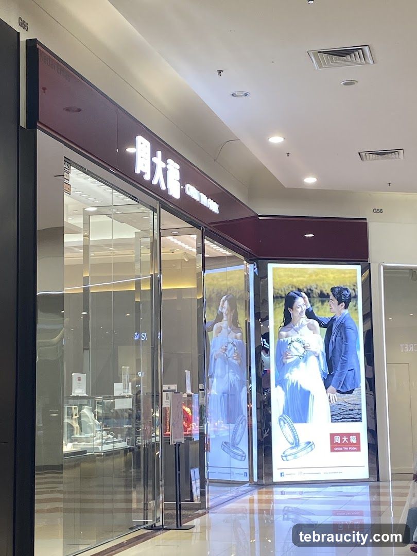The exterior of a Chow Tai Fook jewelry store. The store has a large glass facade with displays of jewelry visible inside. Above the entrance, a prominent illuminated sign displays the store's name in Chinese characters and English. Large vertical advertisements featuring a couple and jewelry are displayed on the right side of the entrance.