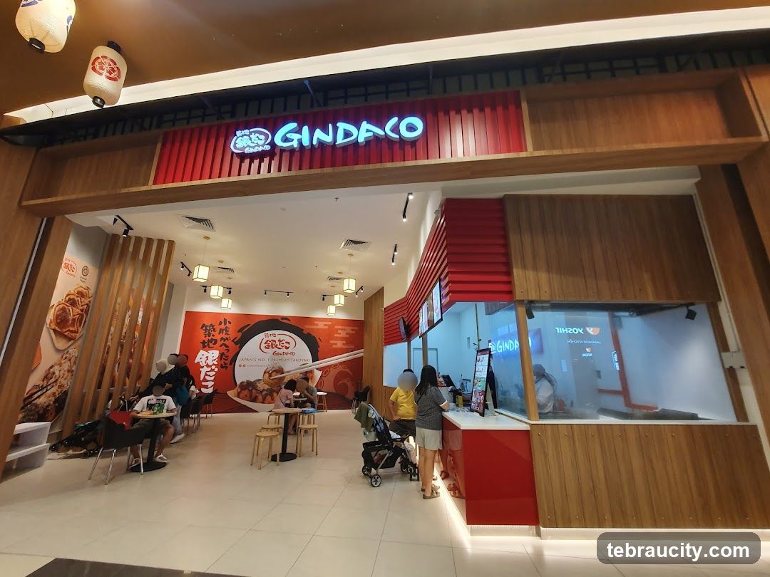 The exterior of a Gindaco restaurant with a prominent sign above the entrance. The restaurant features a modern design with wooden accents and red detailing. Inside, customers are seen dining at tables, and staff are visible behind the counter. Large posters displaying food items are displayed on the walls.