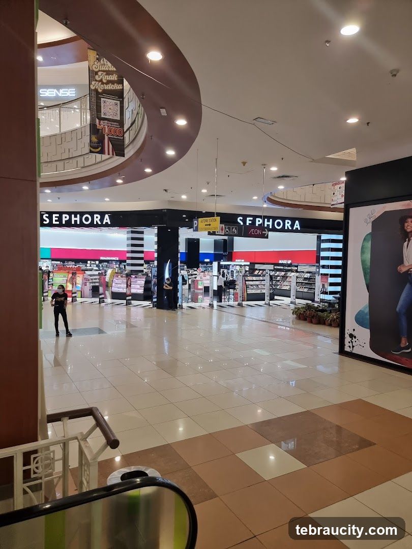 The image shows the exterior of a Sephora store within a mall. The prominent "SEPHORA" logo is visible above the entrance. Inside the store, various makeup and beauty products are displayed on shelves and counters. Several customers and staff members are seen within the store. In the background, other mall shops and signage are visible, including a "SENSE" store and a banner with Malaysian flag elements.