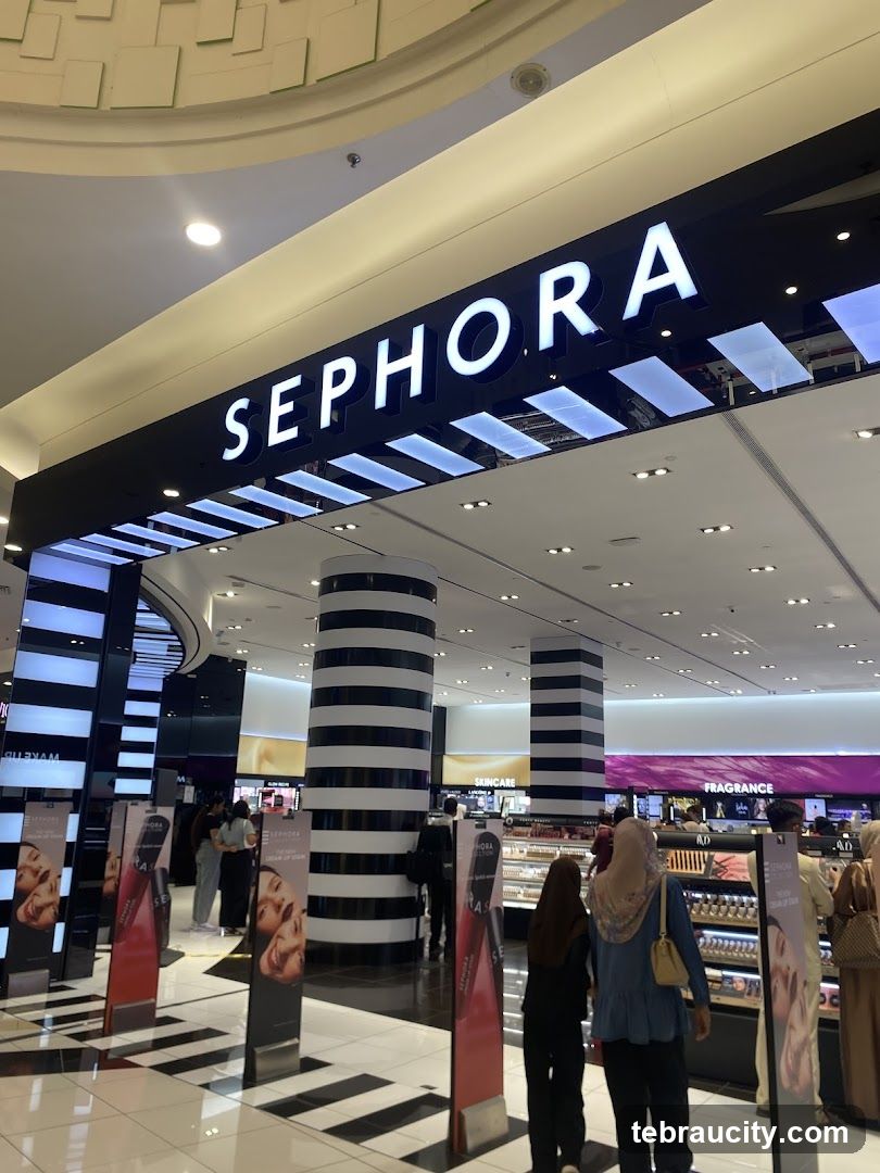 The exterior entrance of a Sephora store with the prominent "SEPHORA" logo illuminated above the entrance. The store features black and white striped columns and displays of beauty products visible inside. Several promotional posters are placed near the entrance.