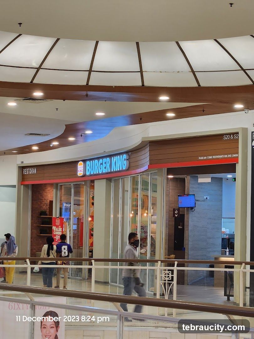 The exterior of a Burger King restaurant located inside a mall. The prominent Burger King logo and name are visible above the entrance. The storefront features large glass windows and doors, with some patrons visible inside. A digital menu board is mounted on the wall to the right of the entrance.