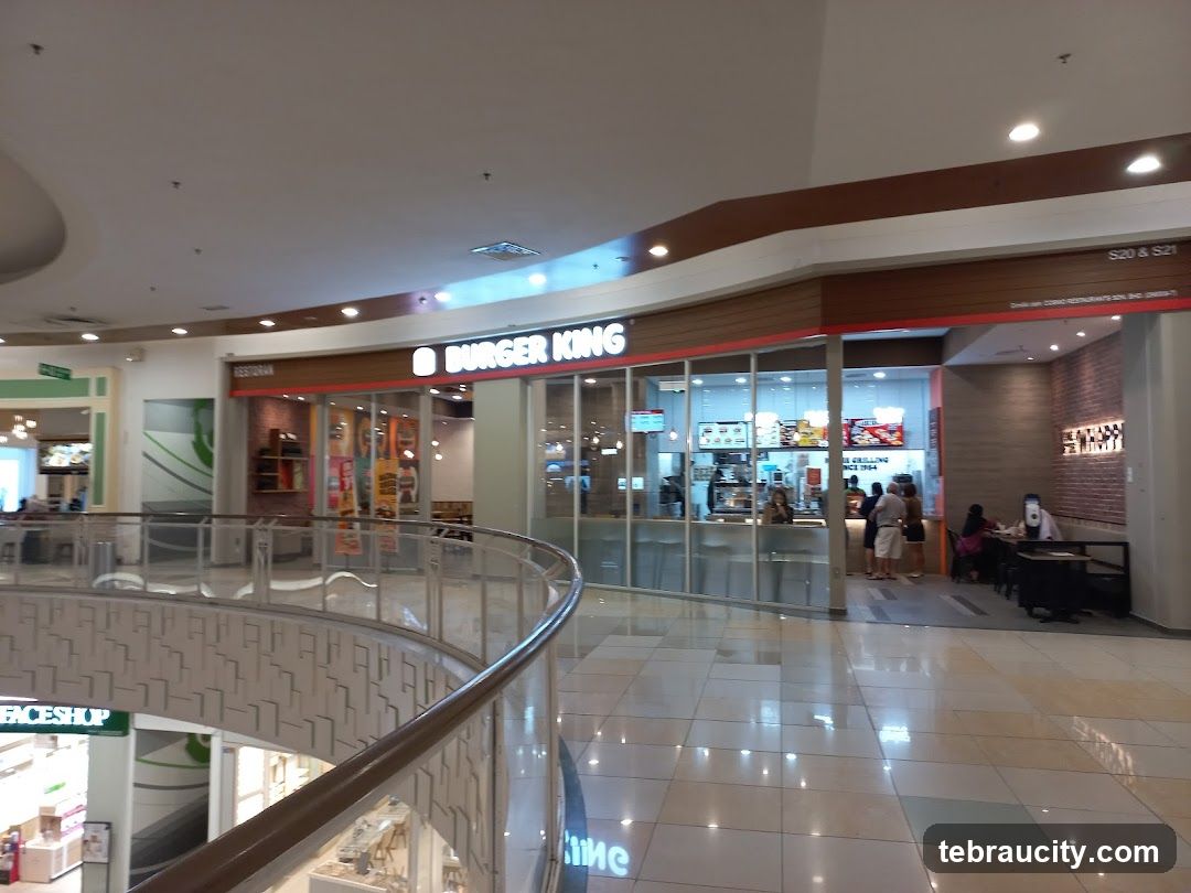 The exterior of a Burger King restaurant located inside a mall. The restaurant has a large glass facade with the Burger King logo and name clearly visible. Inside, customers can be seen ordering and dining.
