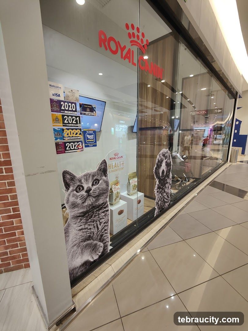 The storefront of a Royal Canin store features large glass windows displaying pet food products and cutouts of a cat and a dog. The prominent red Royal Canin logo is visible above the entrance. Various payment method stickers and year markers (2020-2023) are displayed on the left side of the window. Inside, shelves with pet food bags and boxes are visible.