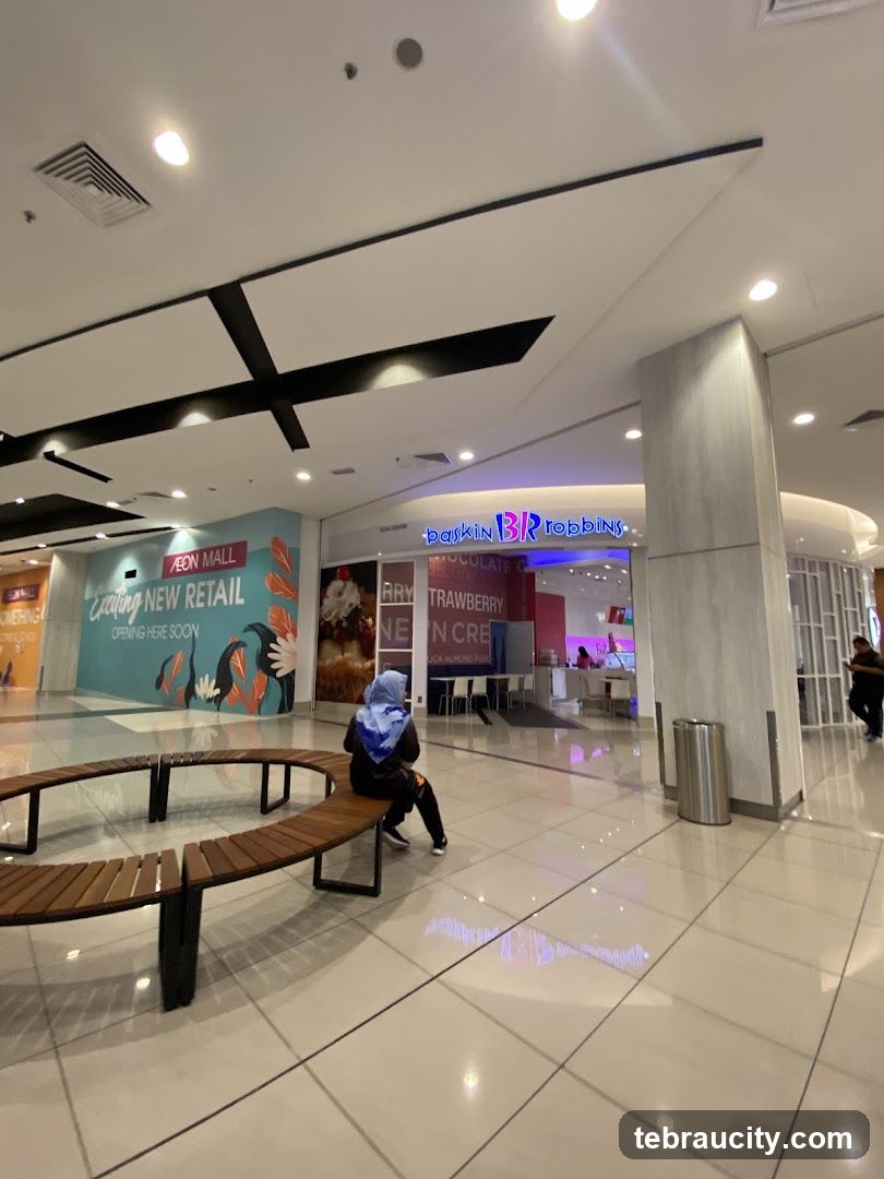 The exterior of a Baskin Robbins ice cream shop in a mall. The store has a bright pink and blue sign with the Baskin Robbins logo. Inside, there are tables and chairs, and staff can be seen behind the counter. To the left of the Baskin Robbins store is a large mural advertising "AEON MALL Exciting New Retail Opening Here Soon." A curved wooden bench is in the foreground.