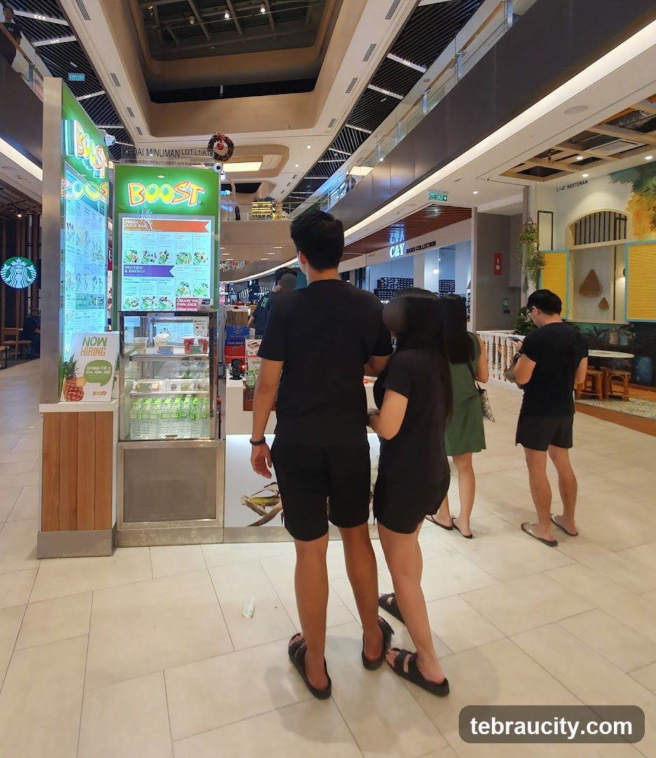 A Boost Juice Bar storefront in a mall. The facade features a large green sign with the Boost logo and menu boards displaying various juice options. A display case with bottled drinks is visible below the menu. A "Now Hiring" sign with a pineapple graphic is also present.