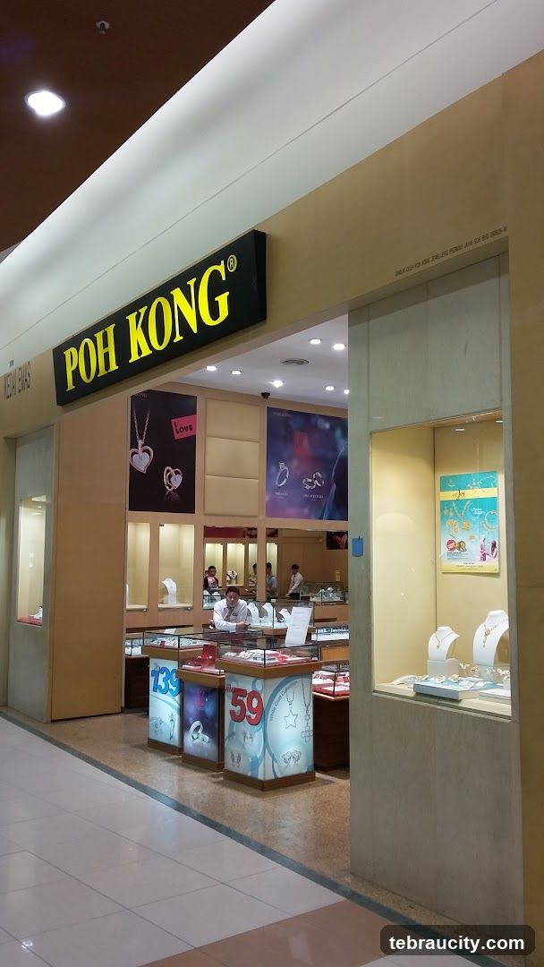 The exterior of a Poh Kong jewelry store. A large yellow sign with black lettering that reads "POH KONG" is visible above the entrance. Inside, display cases showcase jewelry, and staff members are visible. Promotional posters featuring jewelry are displayed on the walls.