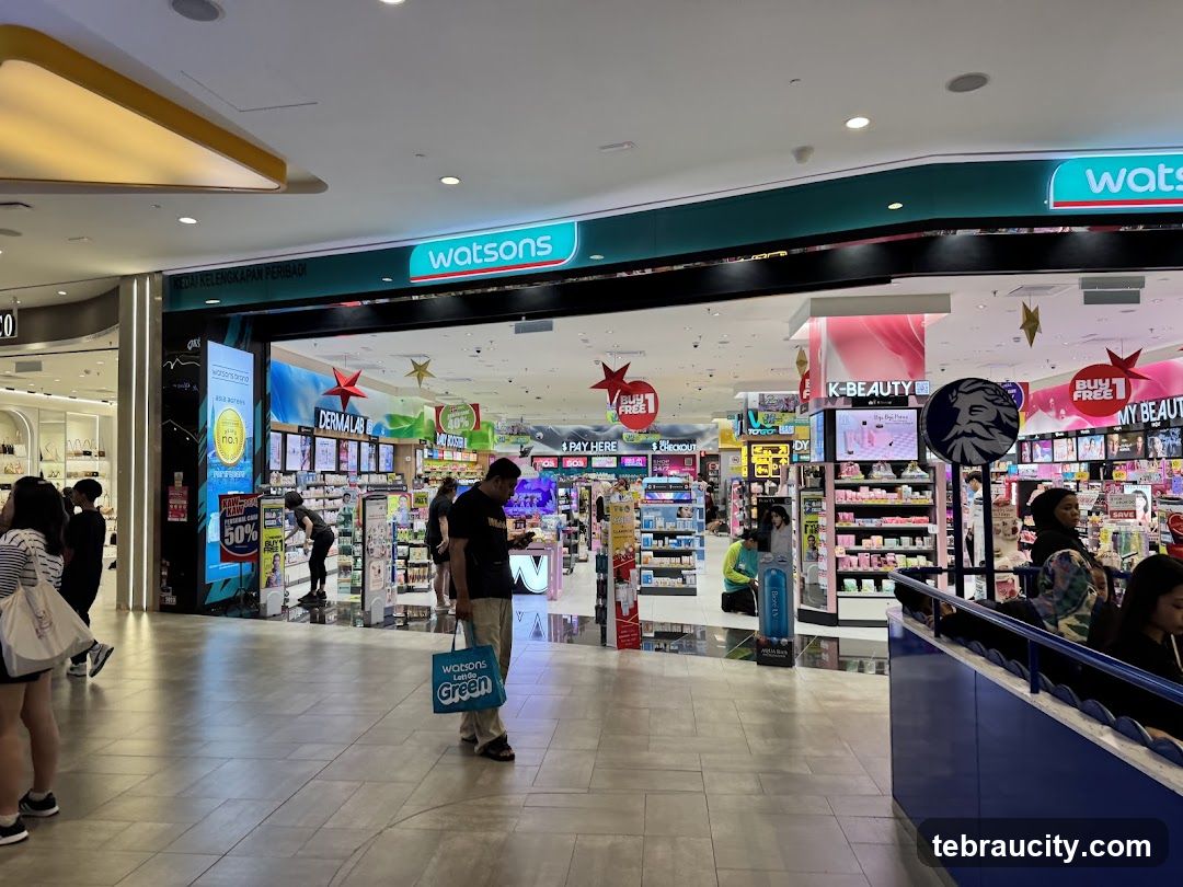 The exterior of a Watsons store, a personal care and beauty retailer, is visible from the inside of a mall. The store features a wide entrance with shelves stocked with various products. Signage for "Watsons" is prominently displayed above the entrance. Promotional signs for "K-BEAUTY" and "BUY 1 FREE 1" are also visible. Several customers are browsing the aisles.