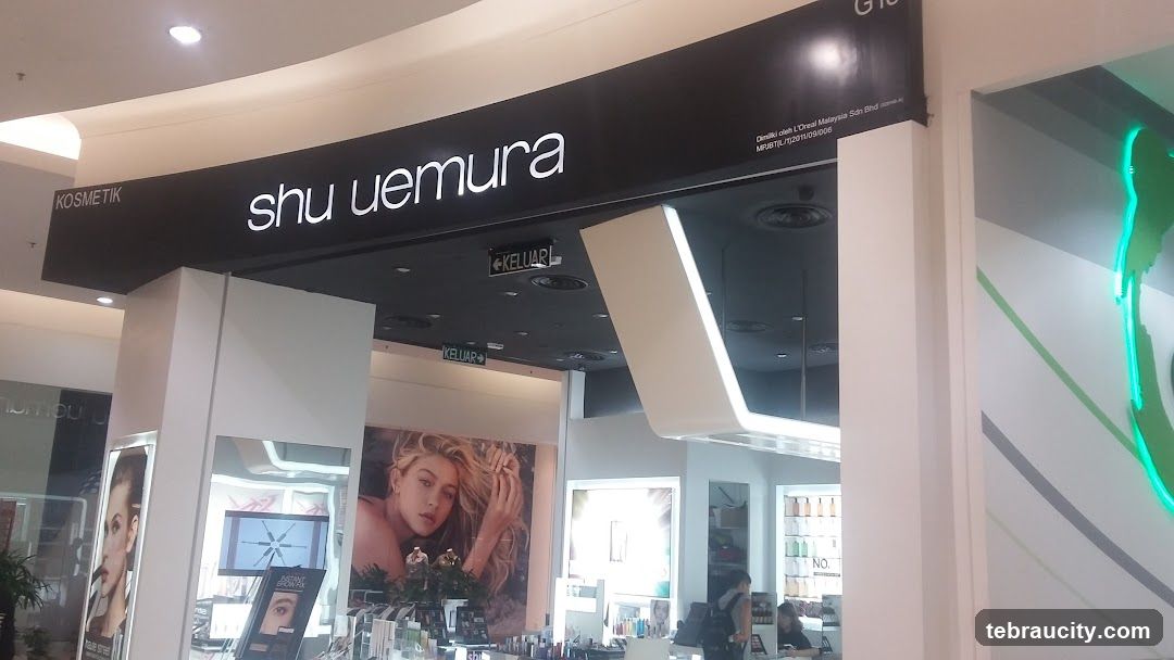 The exterior of a Shu Uemura cosmetics store is visible, with the brand name prominently displayed on a black sign above the entrance. The store features large displays with images of models and cosmetic products. Signs indicating "KELUAR" (Exit) are also visible.