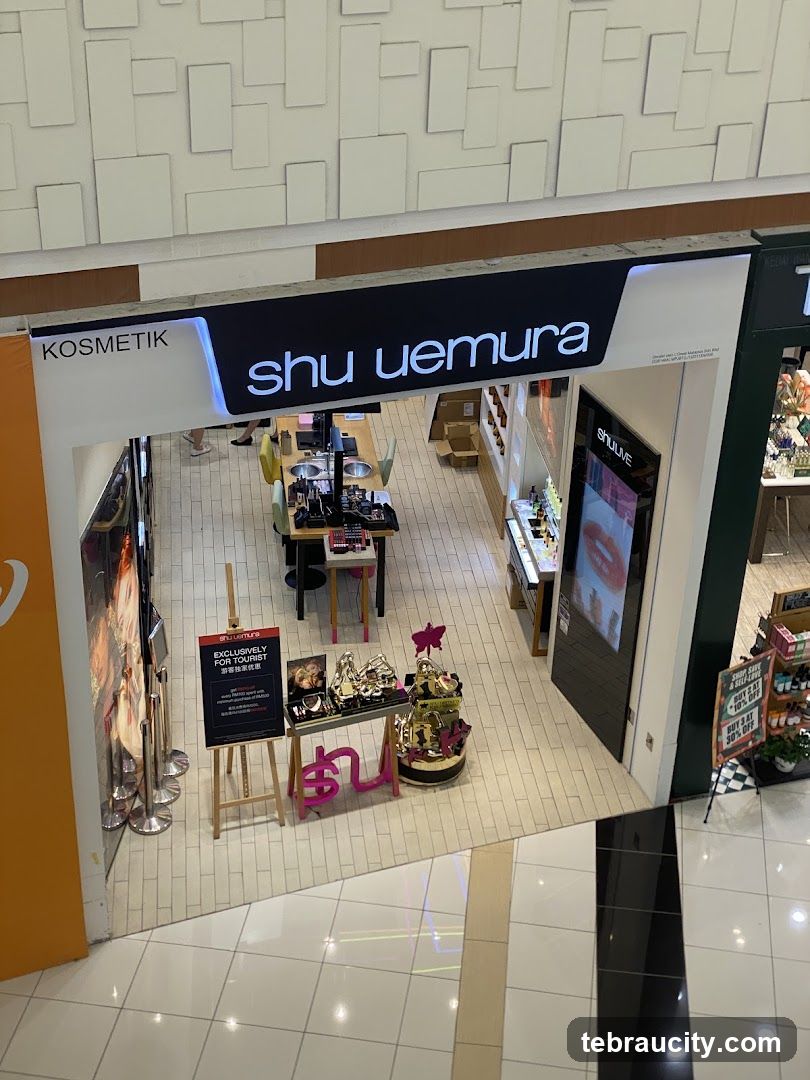 The exterior of a Shu Uemura cosmetics store. The store has a large sign above the entrance that reads "shu uemura" and "KOSMETIK". Inside, makeup products are displayed on tables and shelves. There is also a display stand with promotional materials and products outside the entrance.