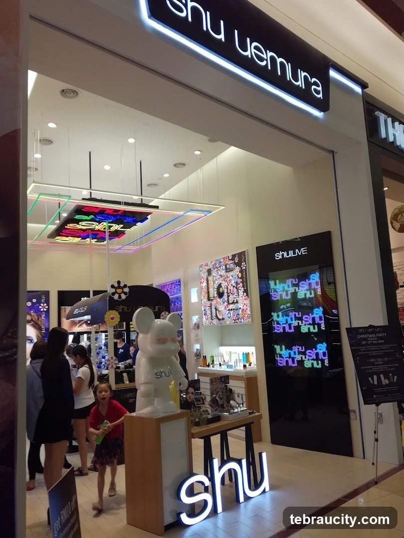 The exterior of a Shu Uemura store with a prominent illuminated sign above the entrance. Inside, various cosmetic products are displayed on shelves and tables. A large white bear-shaped figure with the Shu Uemura logo is positioned near the entrance. Several people are seen browsing inside the store.