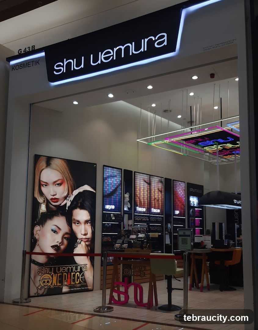The exterior of a Shu Uemura cosmetics store. A large sign above the entrance reads "shu uemura" with "KOSMETIK" to the left. A promotional poster featuring models with dramatic makeup and the Shu Uemura x One Piece collaboration is displayed to the left of the entrance. Inside, shelves are stocked with makeup products, and displays highlight eye, face, and lip products.