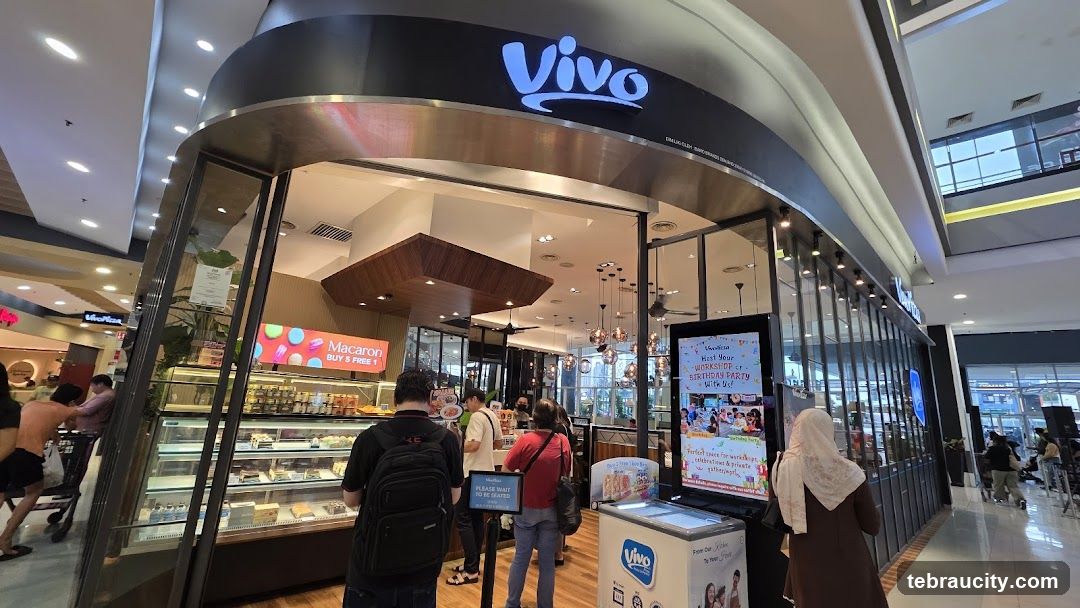 The exterior of a Vivo cafe with a prominent "Vivo" logo above the entrance. Display cases filled with pastries and cakes are visible inside. A sign advertises "Macaron BUY 5 FREE 1". Another display promotes hosting workshops and birthday parties.