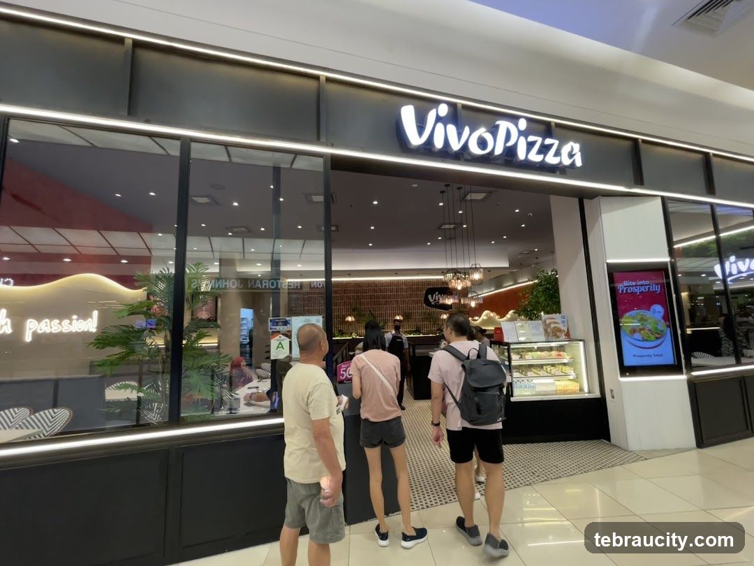 The exterior of a Vivo Pizza restaurant with its prominent signage above the entrance. The storefront features large glass windows, revealing the interior dining area and a display case with food items. A digital advertisement for a "Prosperity Salad" is visible on the right side of the entrance.
