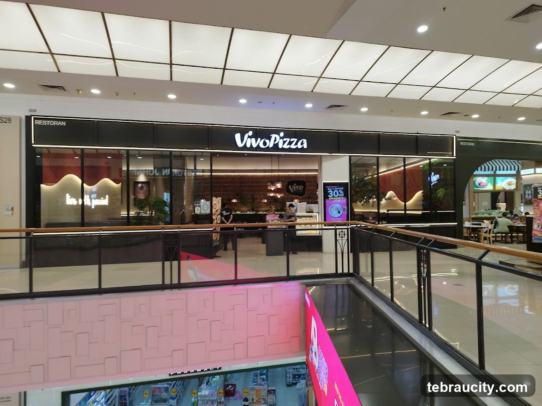 The storefront of Vivo Pizza, a restaurant located in a mall. The name "Vivo Pizza" is prominently displayed in large white letters on a black background above the entrance. The interior of the restaurant is visible through large glass windows, showing seating areas and decor. To the left of Vivo Pizza, there is another establishment with a sign that reads "RESTORAN". To the right, another restaurant with a green awning and visible seating is present. A promotional poster for "Buy 1, Get 1 FREE" is displayed on a digital screen outside the Vivo Pizza entrance.