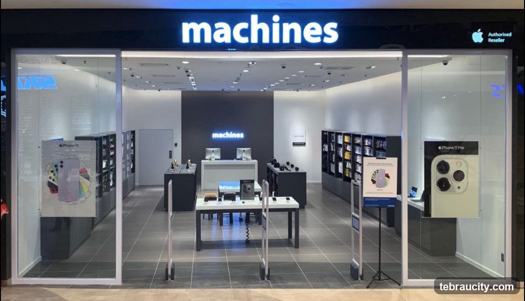 The exterior of a Machines store, an authorized reseller. The store has a modern design with large glass windows and doors. The prominent "machines" logo is displayed in blue neon lights above the entrance. Inside, Apple products are displayed on shelves and tables. Posters advertising the iPhone 11 and iPhone 11 Pro are visible in the windows.