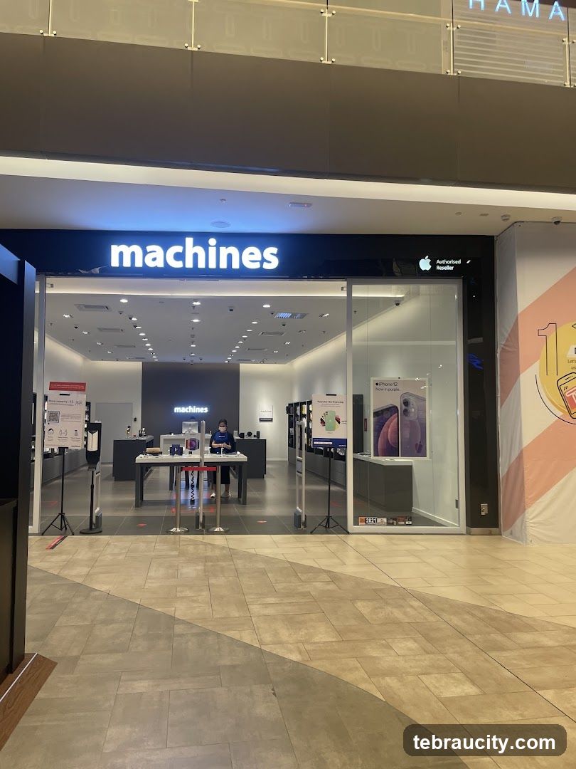 The exterior of a Machines store, an authorized Apple reseller, with its prominent "machines" logo above the entrance. Inside, displays of Apple products are visible, and staff members are present. The store is located within a mall, as indicated by the surrounding architecture and flooring.