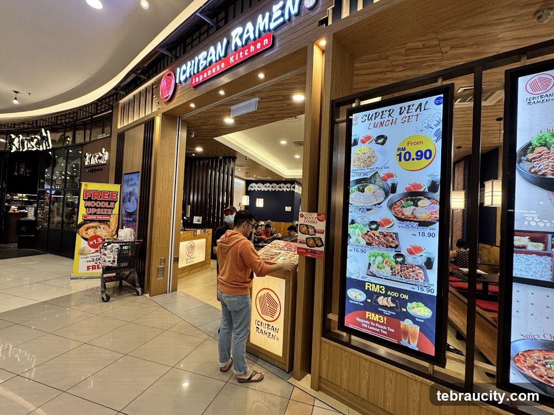 Storefront entrance with illuminated sign displaying 'ICHIBAN RAMEN' and 'Japanese Kitchen'. Wooden paneling frames the doorway. A customer in an orange hoodie stands at the counter, facing inside. The interior shows seated diners and a service counter. Promotional posters and digital menu boards are visible outside but not considered shop_front.