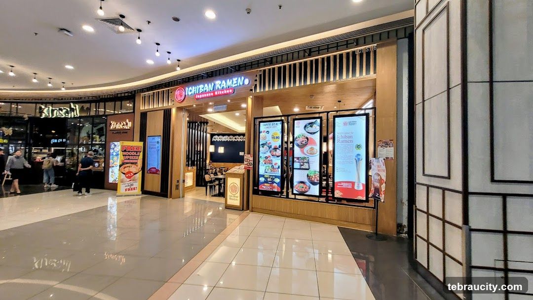 Interior view of a restaurant entrance with illuminated signage reading 'ICHIBAN RAMEN' above the doorway. The facade features wooden paneling and vertical slats. Large digital menu boards display food images and prices. A promotional standee advertises free noodles. Shoppers are visible in the background.