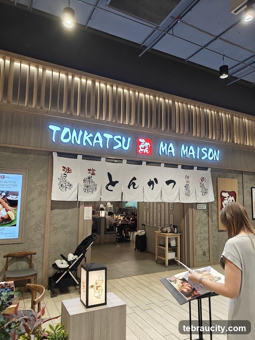 Storefront with illuminated neon sign reading 'TONKATSU MA MAISON' above a traditional Japanese-style entrance curtain featuring calligraphy. The facade includes wooden slat lighting and a textured wall. A stroller is parked outside, and a customer stands nearby holding a menu stand. Inside, diners are seated at tables.