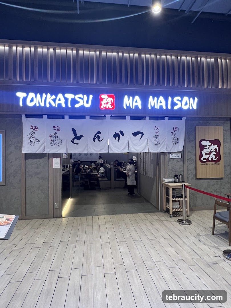 Storefront with illuminated sign reading 'TONKATSU MA MAISON' above entrance. White fabric banner with Japanese characters hangs across doorway. Interior shows patrons dining and staff working. Modern lighting and wood-look flooring. Red velvet rope barrier to right of entrance.