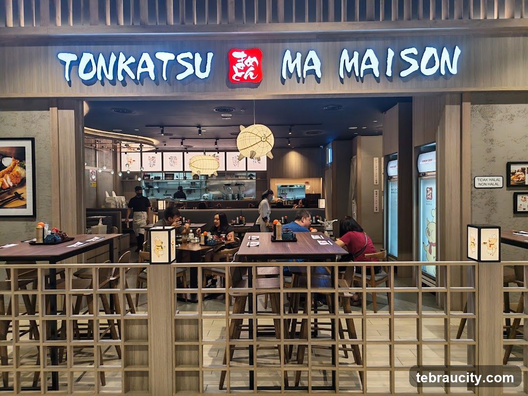 Exterior view of restaurant entrance with illuminated sign reading 'TONKATSU MA MAISON' above the doorway. The facade features wooden paneling and a decorative lattice barrier in front. Inside, patrons are seated at tables, staff are visible behind counter, and hanging lanterns illuminate the interior.