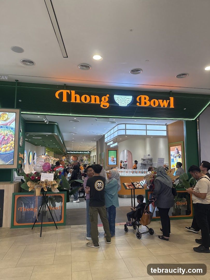 Storefront with illuminated orange signage reading 'Thong Bowl' above a green facade; customers queue inside the entrance; promotional floral arrangement on stand to left; interior shows bright lighting and modern design.