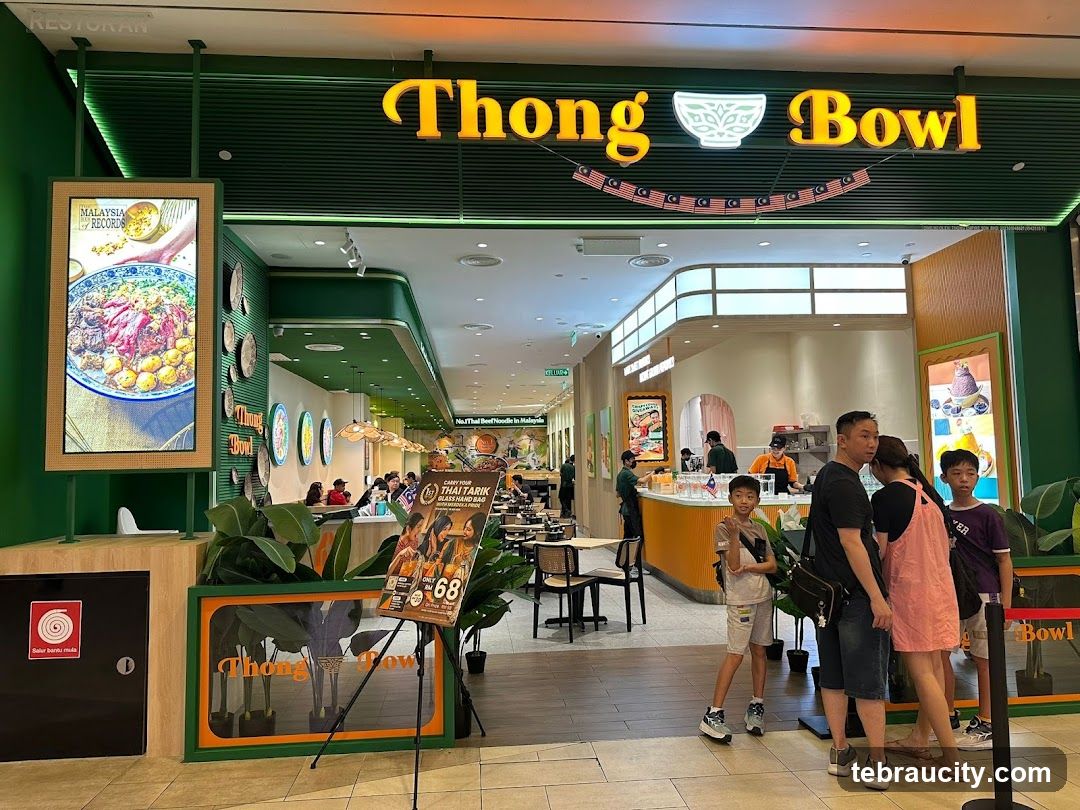 Green-themed restaurant entrance with illuminated 'Thong Bowl' sign featuring a bowl logo. A Malaysian flag banner hangs below the sign. Customers are seen near the counter area inside. Large digital screens display food imagery on either side of the entrance. The interior shows seated diners and staff behind the counter.