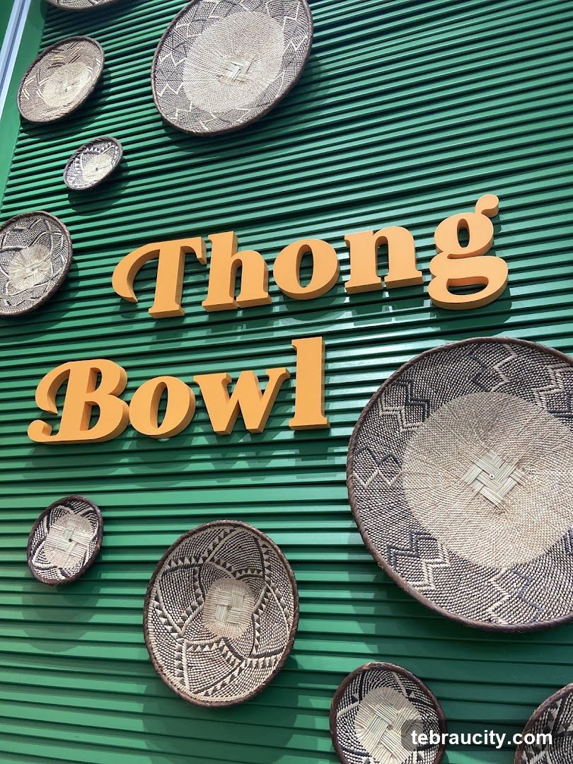 Green corrugated wall with orange 3D lettering spelling 'Thong Bowl'. Multiple woven baskets of varying sizes are mounted on the wall as decorative elements.