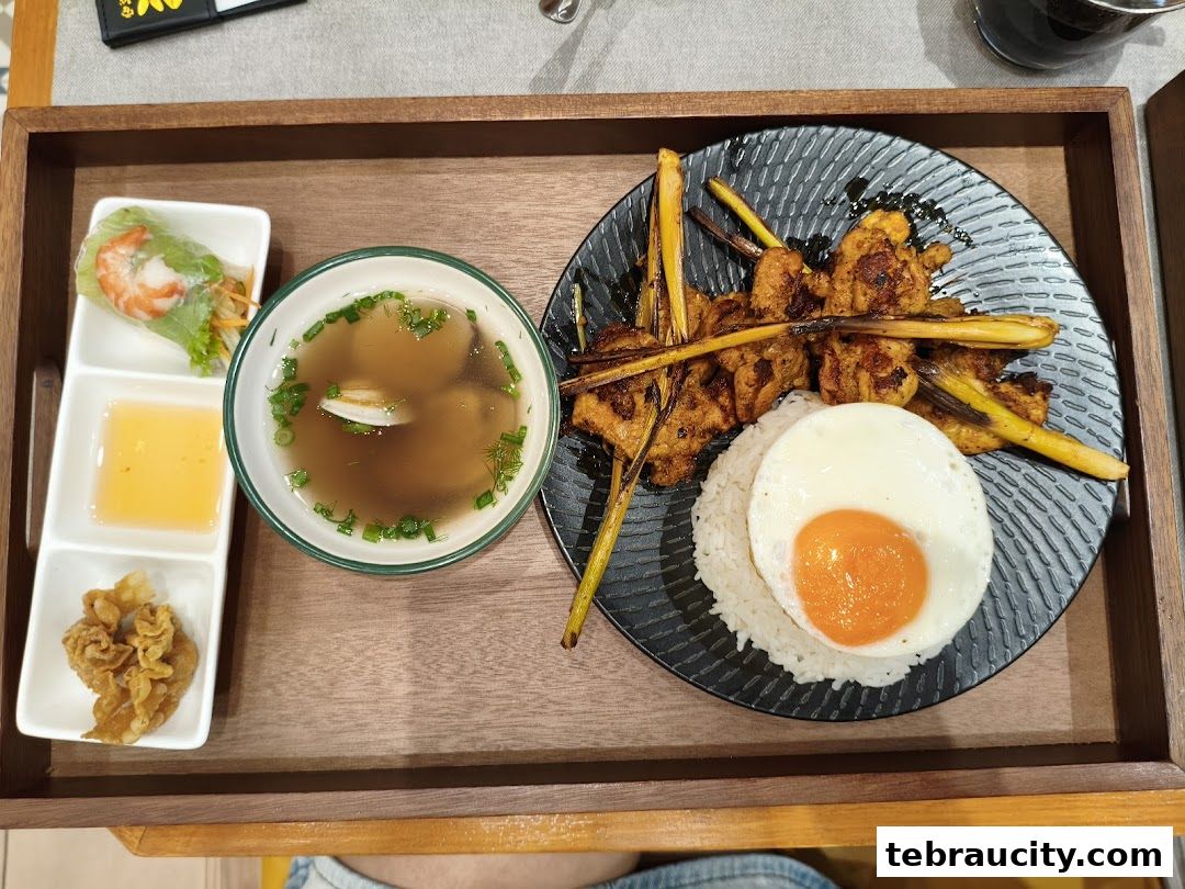 A Vietnamese meal featuring lemongrass chicken skewers, rice, a fried egg, spring roll, and soup.