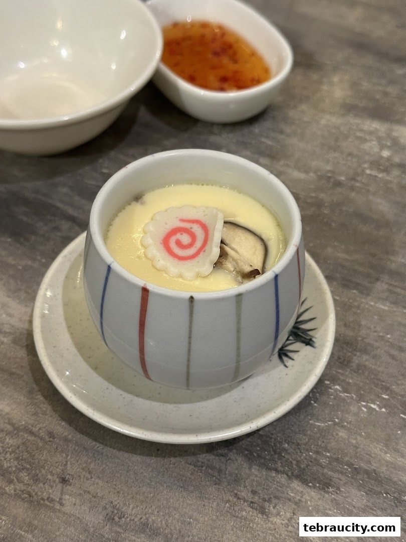 A close-up of a delicate steamed egg custard dish with a decorative fish cake and mushroom.