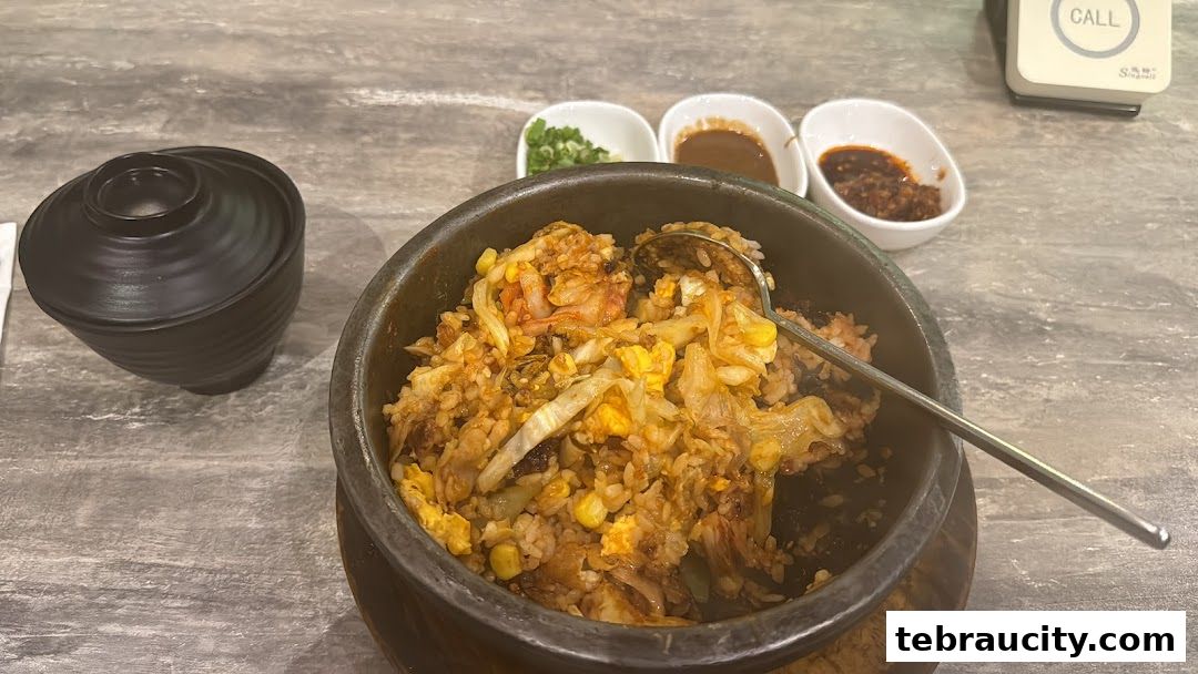A close-up of a steaming hot bibimbap dish with kimchi, corn, and egg, served with dipping sauces.