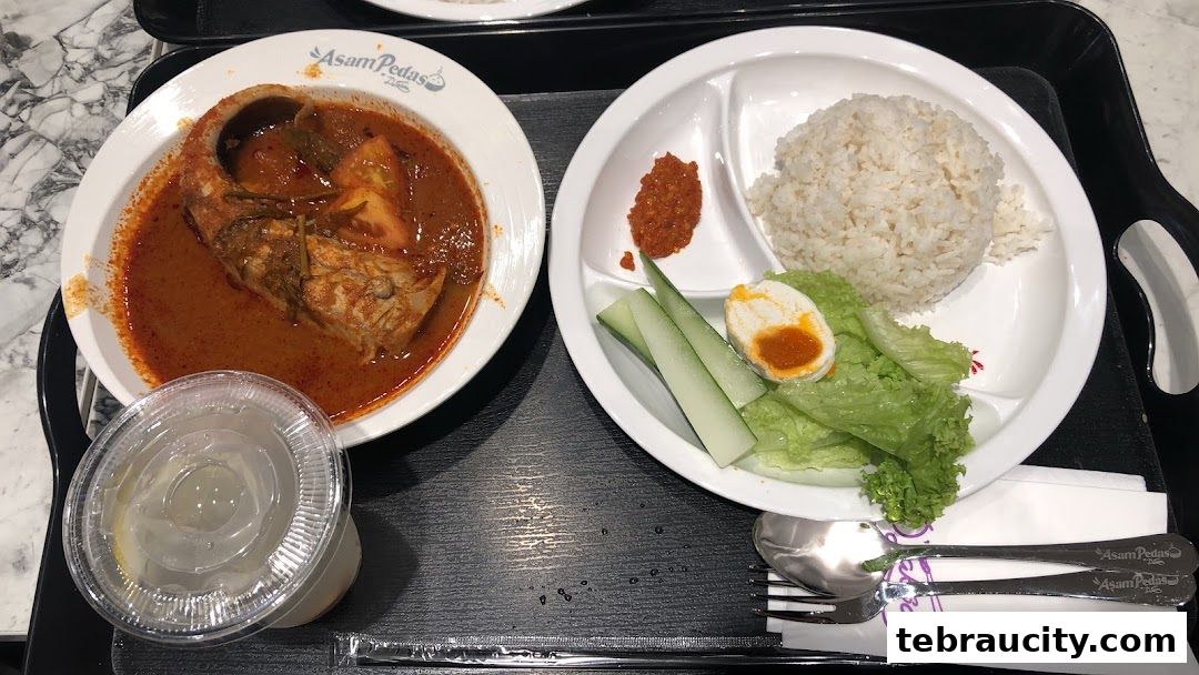 A tray of Asam Pedas fish curry with rice, vegetables, and a drink.
