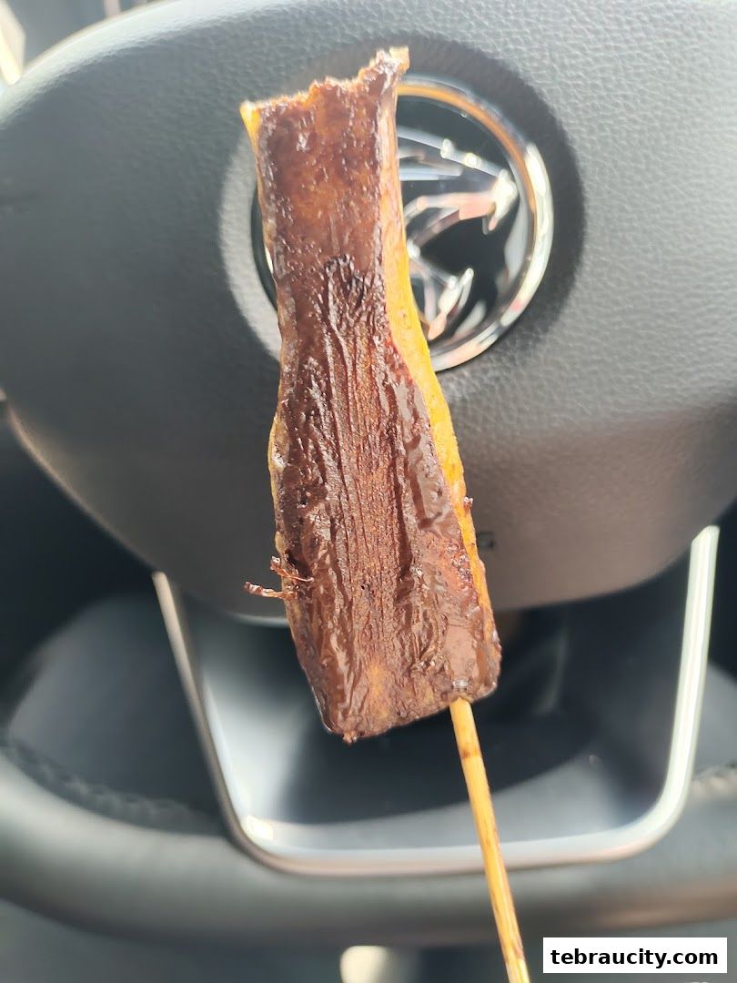 A close-up of a pretzel stick coated in chocolate, held in front of a car's steering wheel.