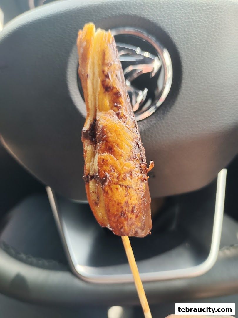 A close-up of a chocolate-dipped pretzel on a stick, held in front of a car's steering wheel.