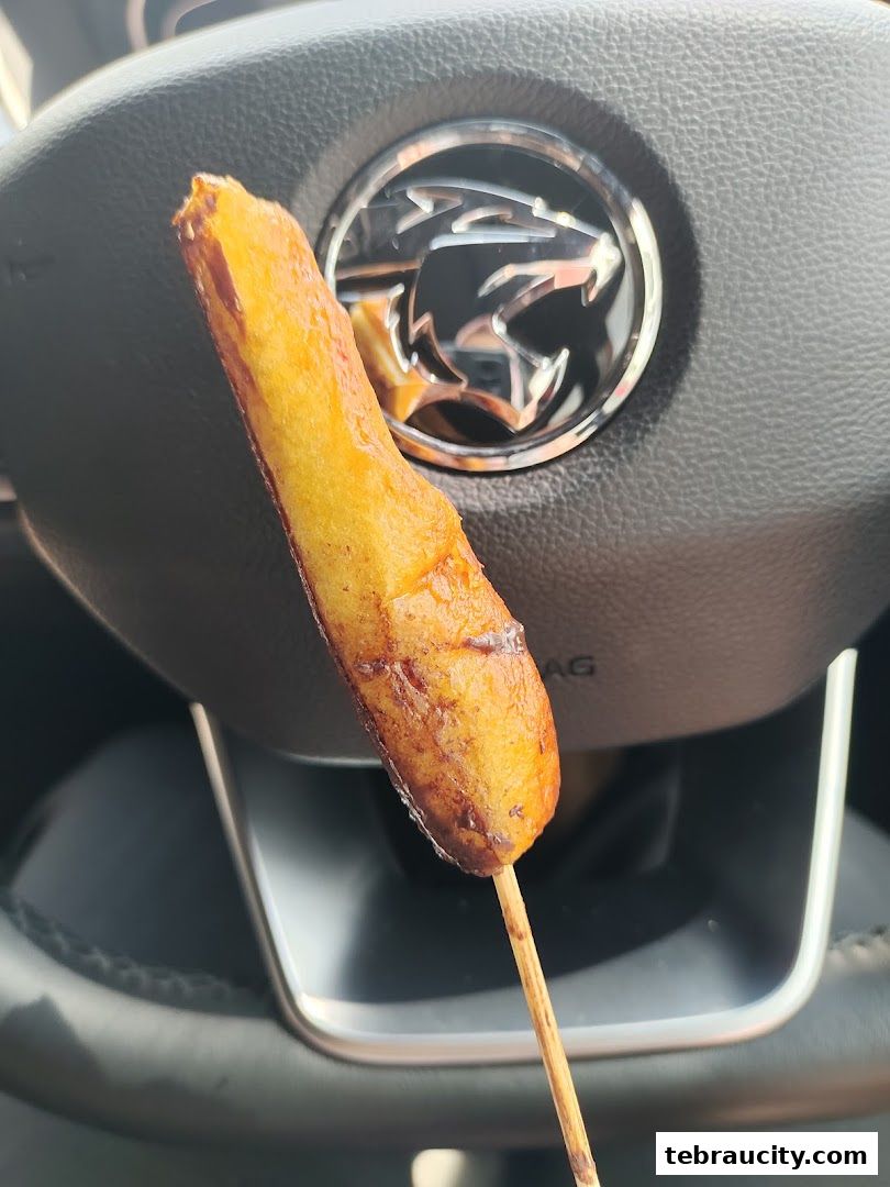 A golden-brown pretzel stick on a wooden skewer in front of a car's steering wheel.