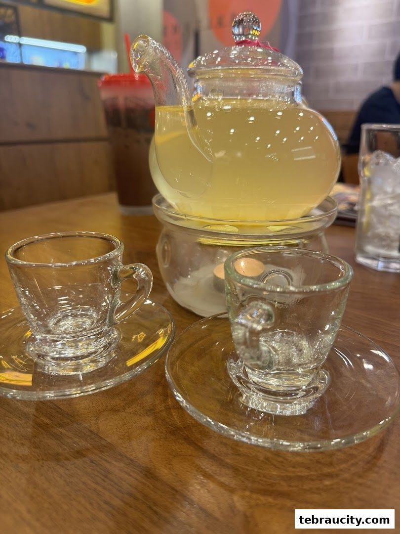 A glass teapot filled with a yellow beverage sits on a warmer with two empty teacups and saucers.