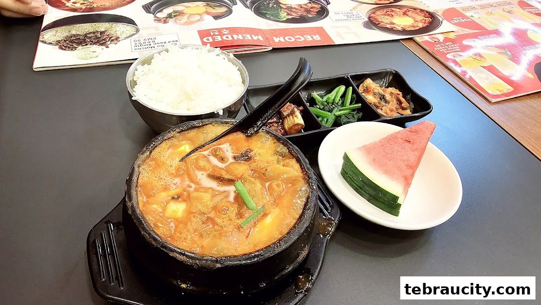 A steaming bowl of Korean stew with rice, kimchi, and a slice of watermelon.