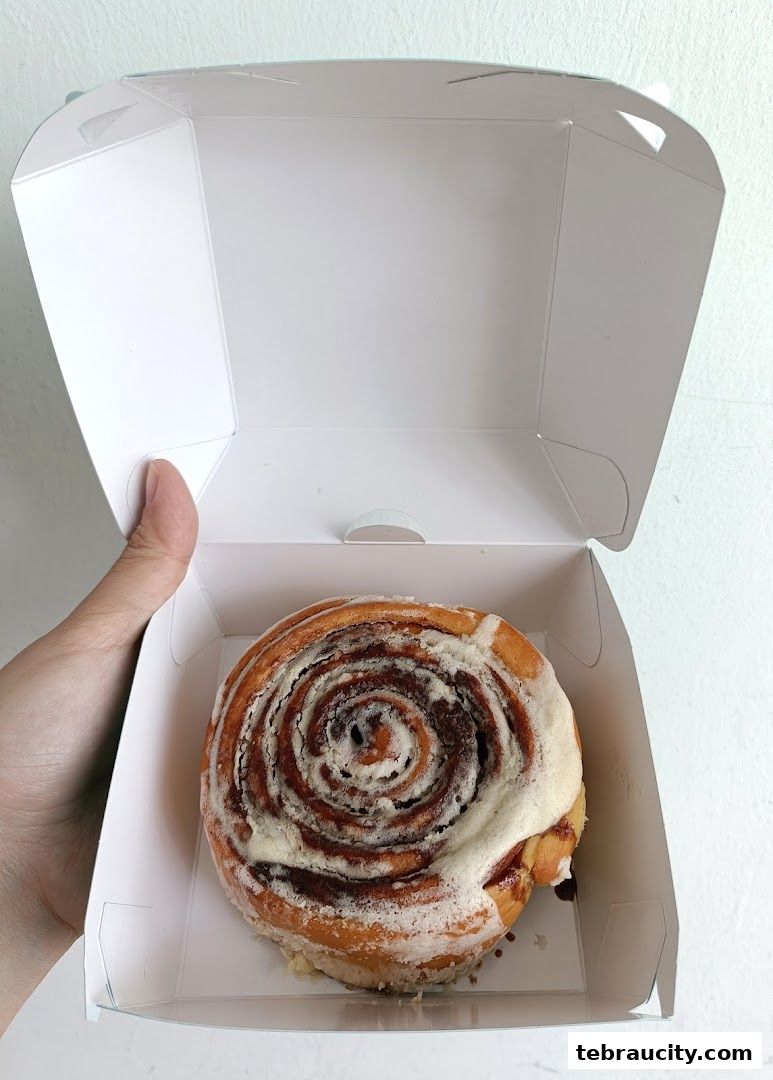 A hand holds open a white box revealing a freshly baked cinnamon roll.