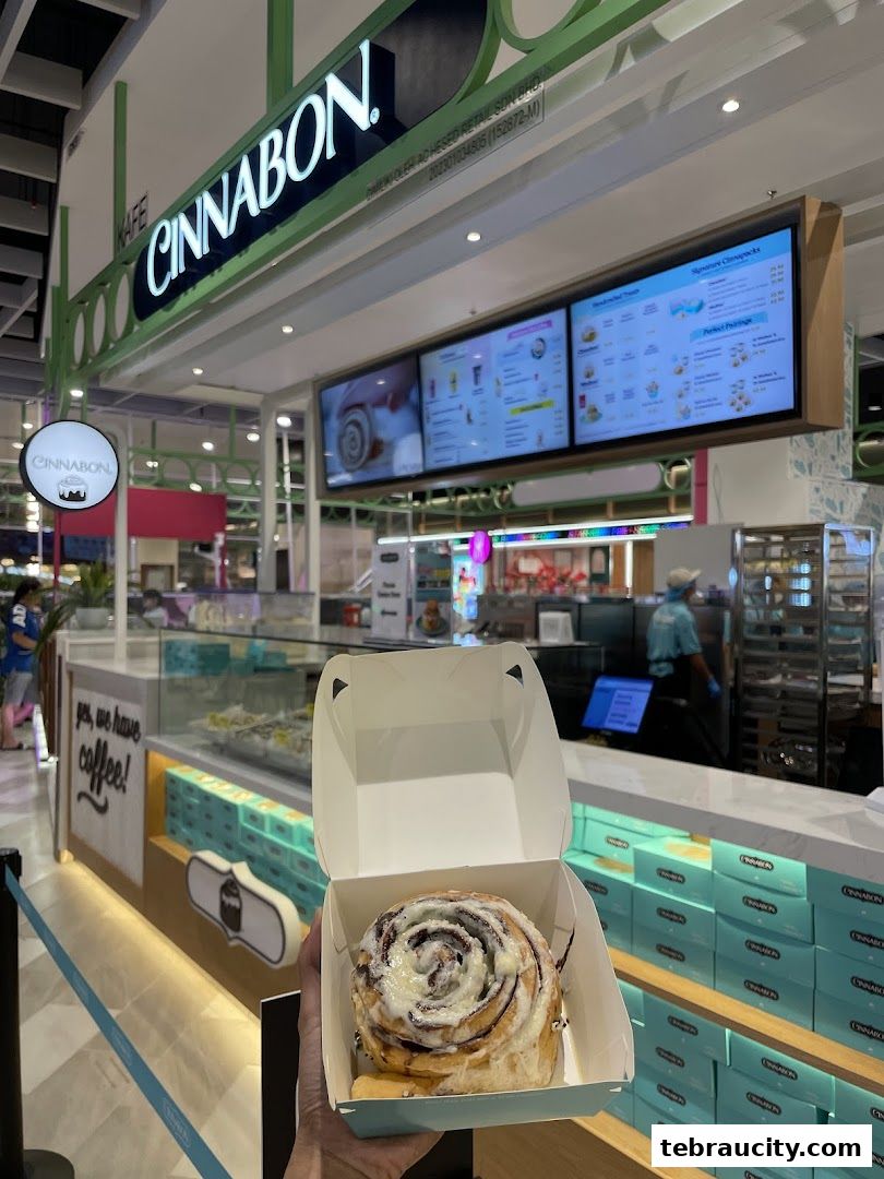 A hand holds a Cinnabon cinnamon roll in a box in front of the Cinnabon store.