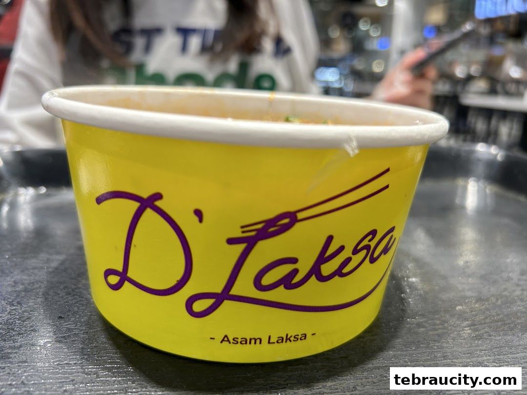 A close-up of a yellow cup with the D'Laksa logo and 'Asam Laksa' text.