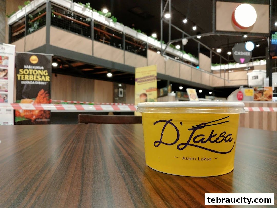 A yellow container with the D'Laksa logo sits on a wooden table in a food court.