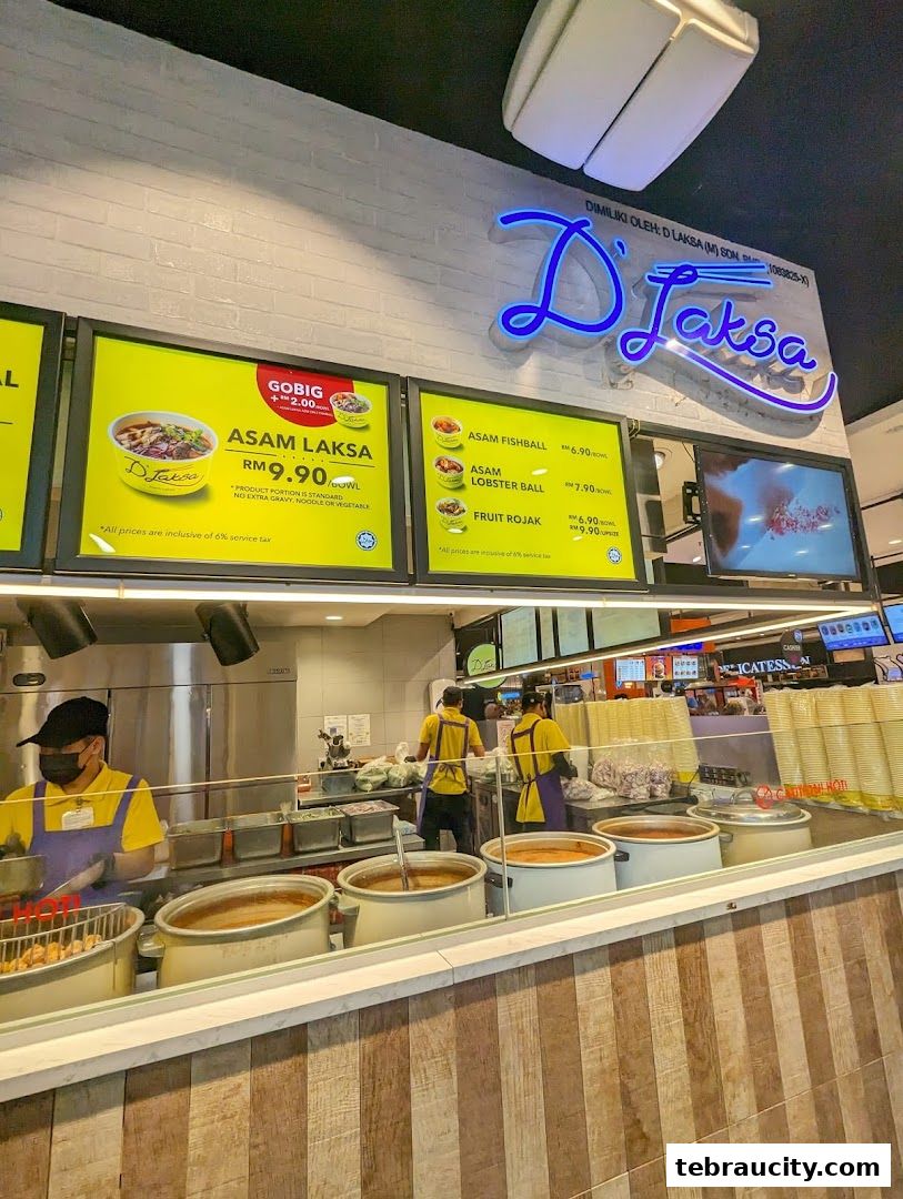 A food stall counter with staff preparing dishes and a menu board displaying Laksa and other items.