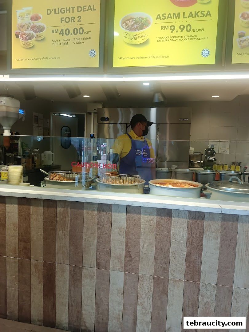 A food stall counter with a staff member and promotional signs for laksa deals.
