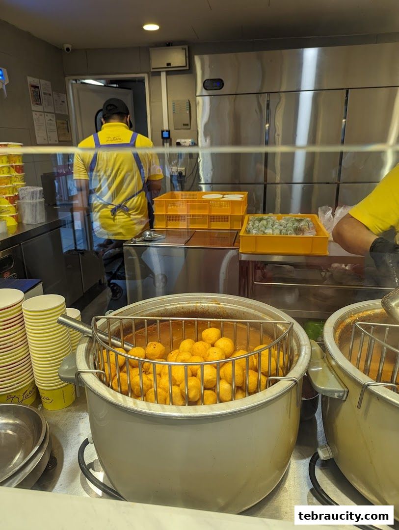 A shop worker in a yellow uniform prepares food behind a counter with simmering pots.