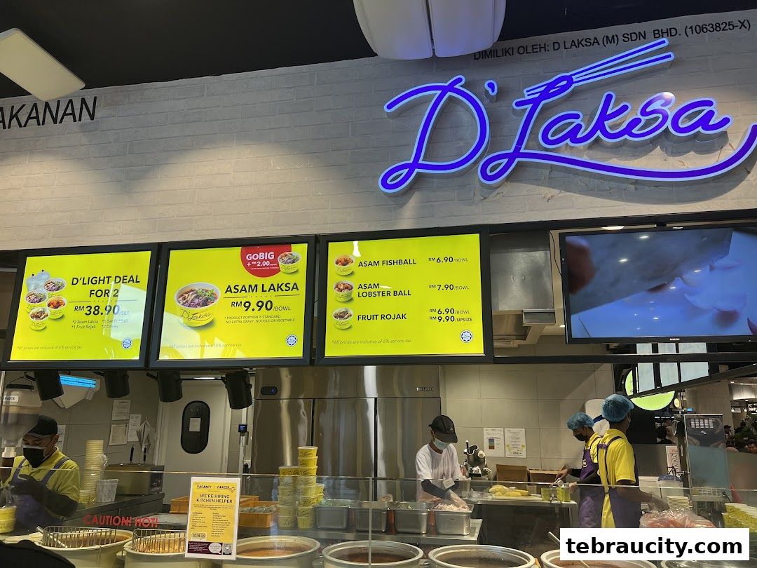 A food stall with a prominent D'Laksa sign and illuminated menu boards displaying various dishes and prices.