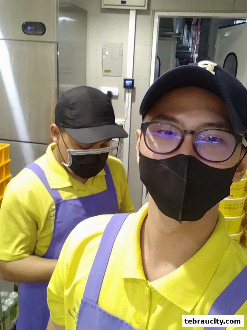 Two staff members wearing yellow shirts and purple aprons pose for a selfie in a kitchen.
