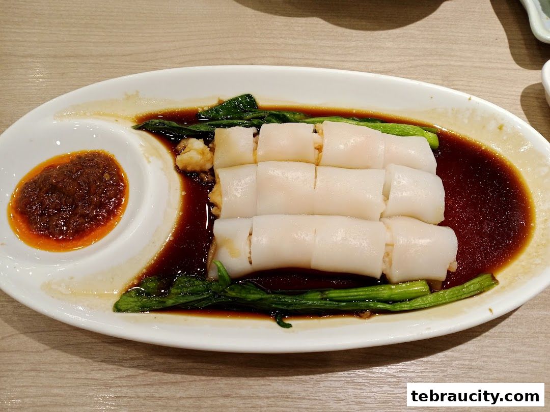 A plate of dim sum chee cheong fun with vegetables and chili sauce.
