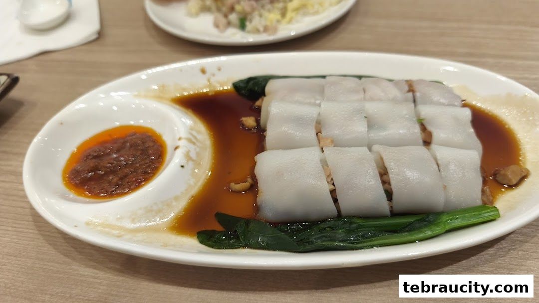 A plate of dim sum chee cheong fun with a side of chili paste and greens.