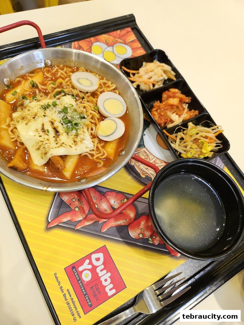 A delicious Korean meal featuring ramen, tteokbokki, eggs, and side dishes on a branded tray.